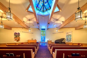 Indoor religious area of worship with stained glass window and skylight, the room has several rows of pews and a piano